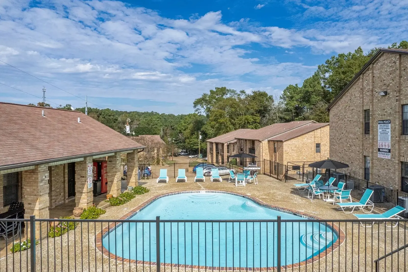 Fenced swimming pool surrounded by lounge chairs and brick apartment buildings.